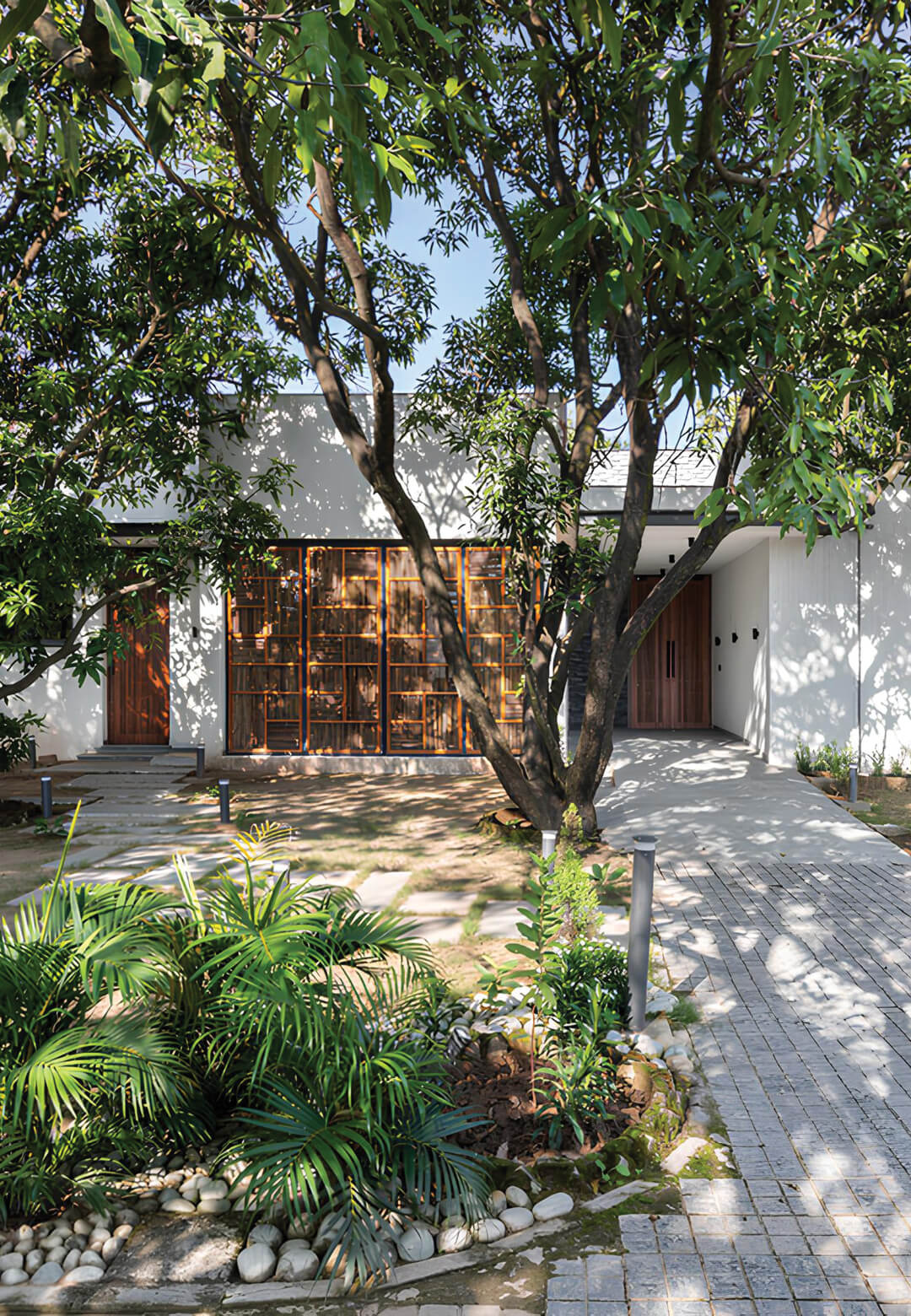The holiday home entrance is framed by lush, mature trees, creating a natural canopy that casts dappled shadows on the paved pathway | House of Mango Shadows by Design i.O | STIRworld